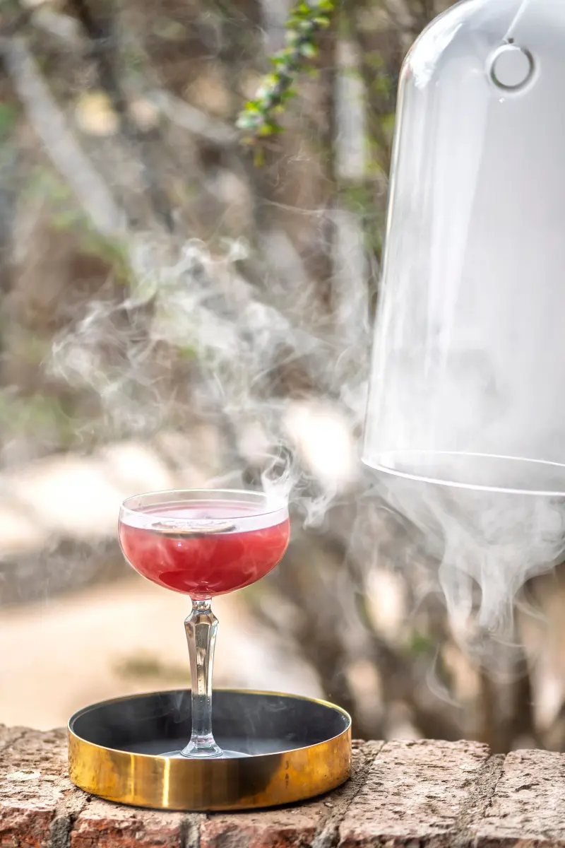 Smoked craft cocktail revealed from under a glass cloche on a restaurant patio